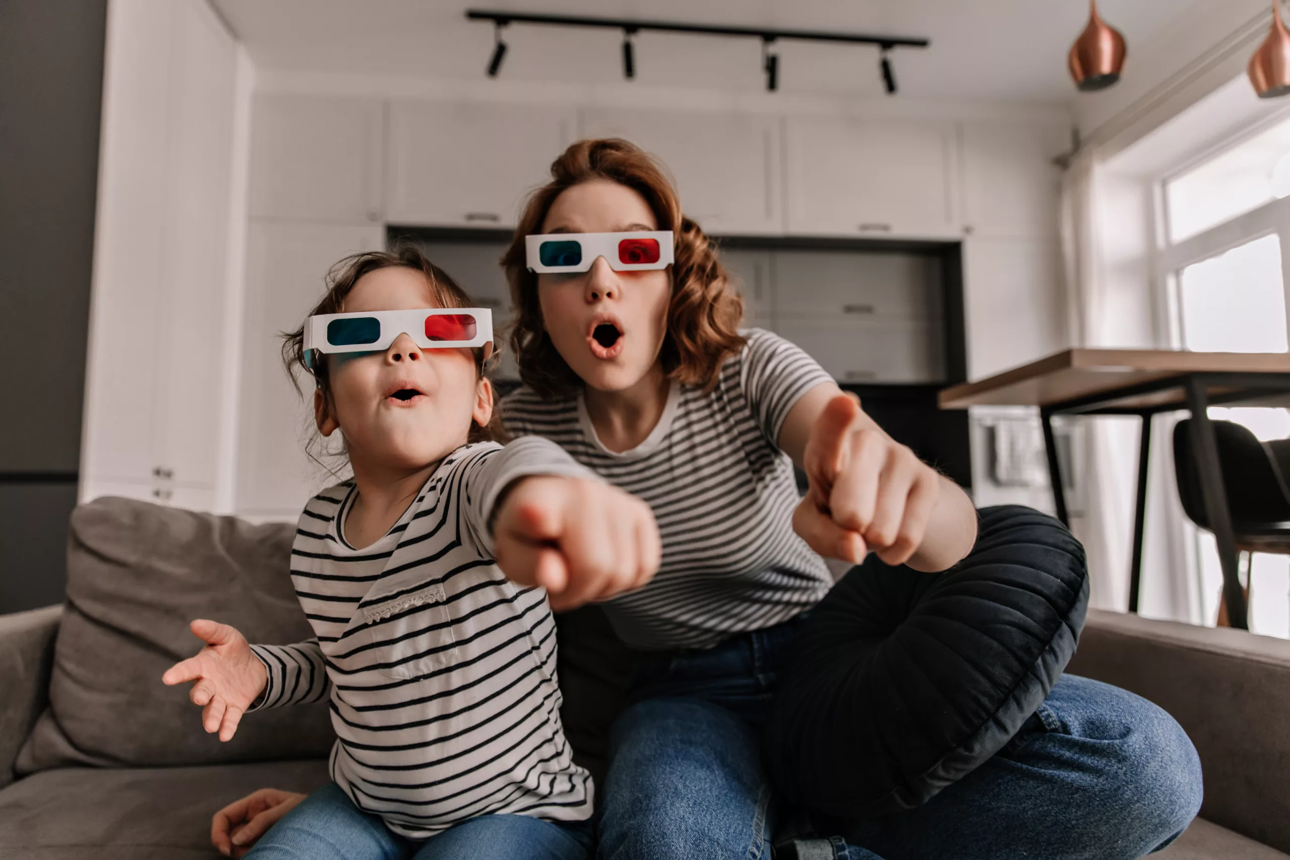 Curly woman and her daughter are sitting on sofa in 3D glasses and are watching movie with surprise